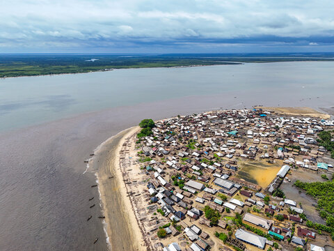 Aerial view of a dense coastal settlement with numerous small houses, a sandy beach lined with fishing boats, and the confluence of a river and the Atlantic Ocean in Monrovia, Liberia.