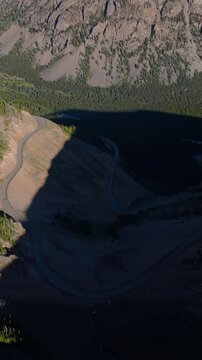 Vertical Screen: Beartooth Highway winds through dramatic Montana USA mountain landscape with steep rocky cliffs and dense evergreen forest. Drone captures serpentine road curves.