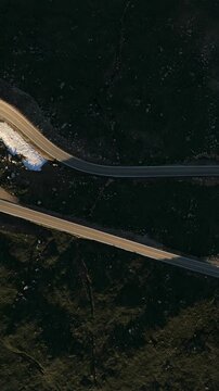 Vertical Screen: Beartooth Highway in Montana USA captured by drone showing winding mountain road with yellow center lines cutting through dark alpine terrain with patches of melting snow.