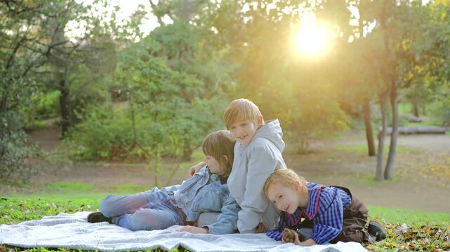 Three siblings relax on a white picnic blanket in a scenic park setting with beautiful golden hour sunlight streaming through lush green trees, creating a warm and peaceful family moment outdoors.