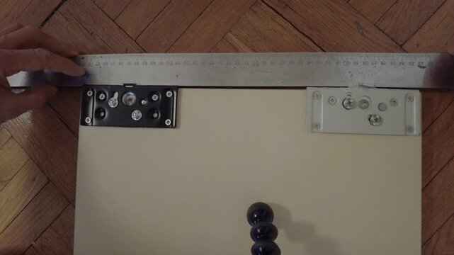 Ruler measuring roller brackets on panel top, hands holding metal ruler across door panel checking alignment of bottom roller assemblies on parquet floor, closeup inspection