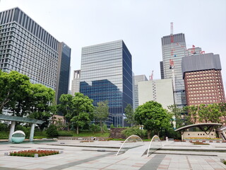 現代都市の広場が広がる景観。ガラス張りの高層ビル群と建設中のクレーンが立ち並び、緑地と歩行者が点在する。 © BRUCE