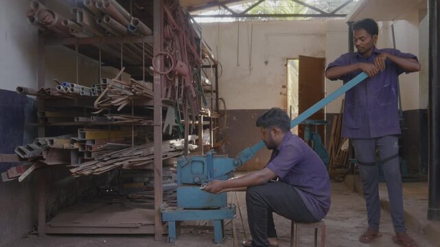 A close indoor shot of two men or male skilled workers or craftsmen, working together, precisely cutting a metal rod into different shapes using a manual cutting machine in a fabrication workshop
