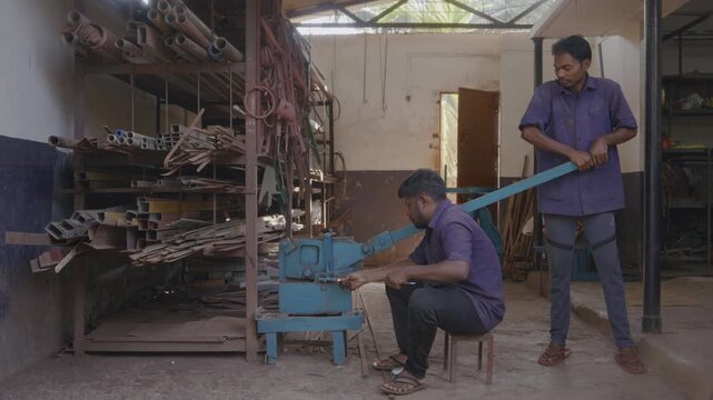 A close indoor wide shot of two men or male skilled workers or craftsmen, working together, precisely cutting a metal rod into different shapes using a manual cutting machine in a fabrication workshop
