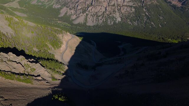 Aerial view of Beartooth Highway winding through Montana mountains with dramatic shadows cast across valley floor, lush green pine forests covering steep slopes.