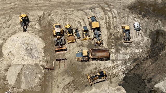 Aerial view of heavy construction machinery including excavators, dump trucks, and graders parked on a sandy site in the United States.