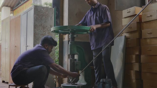 A wide indoor shot of two Indian Asian male skilled workers or craftsmen precisely bending a metal rod into shape using a manual bending machine in a fabrication workshop.