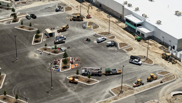Aerial view of a construction site featuring heavy machinery, excavators, rollers, and various utility trucks parked on a paved lot near a large warehouse building in the United States.