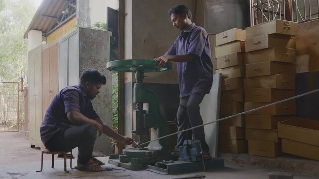 A wide indoor shot of two Indian Asian male skilled workers or craftsmen precisely bending a metal rod into shape using a manual bending machine in a fabrication workshop. 