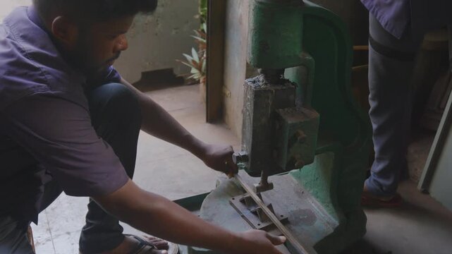 A close up indoor shot of two Indian Asian male skilled workers or craftsmen working together, precisely bending a metal rod into shape using a manual bending machine in a fabrication workshop. 
