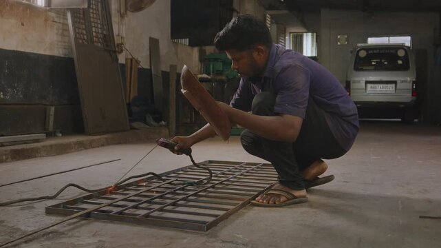 A skilled Indian Asian worker performs arc welding on a custom steel gate, squatting on floor while wearing handheld shield. Sparks fly as the craftsman joins metal components inside fabrication unit