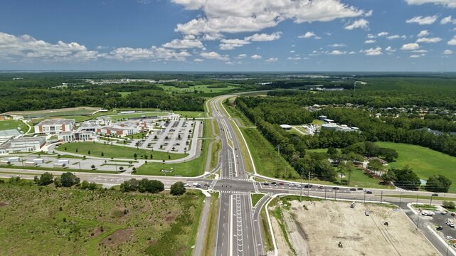 Aerial view of a highway intersection adjacent to a school campus with parking lots, surrounded by lush green forests and open fields under a bright blue sky in the United States.