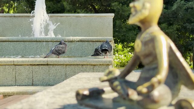 A small bee figurine sits by pigeons resting at a splashing fountain in Simferopol, Crimea. The peaceful sunny park creates a joyful and relaxed feeling.
