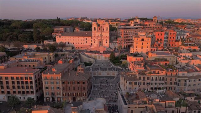 Aerial drone shot in Rome Italy captures the iconic Spanish Steps and Trinita dei Monti Church bathed in warm golden hour light with historic terracotta rooftops and tourists gathering below.