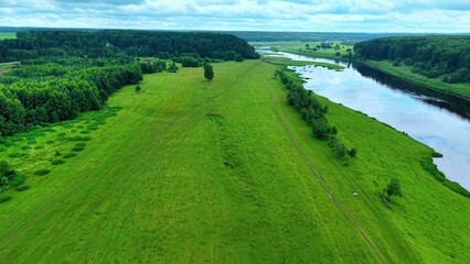 Obraz premium Lush green grasslands bordered by a winding river under a cloudy sky during summer season