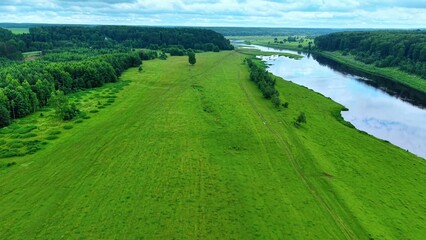 Obraz premium Vast green meadow with flowing river and lush tree line under cloudy sky in summer season