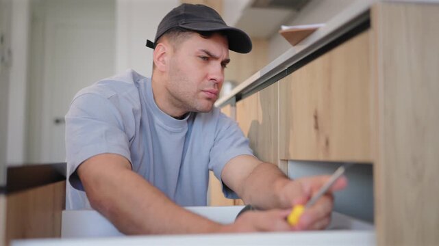 Professional maintenance technician with toolbox in kitchen of home for repair service. Man wearing cap uses screwdriver to fix wooden cabinetry. Handyman focused on detailed work during renovation.