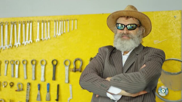Portrait of a confident smiling senior man with a white beard wearing a suit, hat, and sunglasses crossing his arms in a bright workshop. Quirky boss or eccentric mechanic standing by tools.