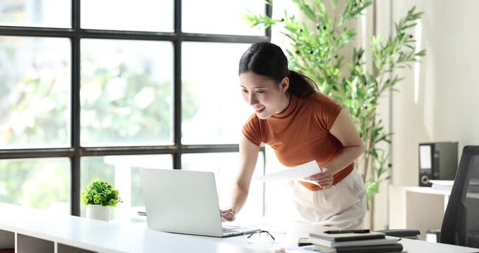 Asian businesswoman smiling and typing on laptop, happily working from a modern home office, staying connected and productive with technology for success