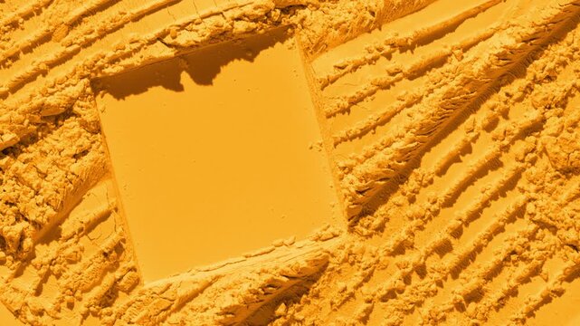 From above, a marigold powder surface showcases intersecting furrows and a square imprint, lit by a light source that projects changing shadows