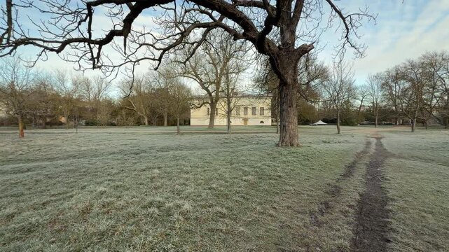A yellow historic mansion sits behind a wide field of silver frost and bare winter trees. This quiet morning scene feels dignified and nostalgic as the soft light hits the cold garden grounds.