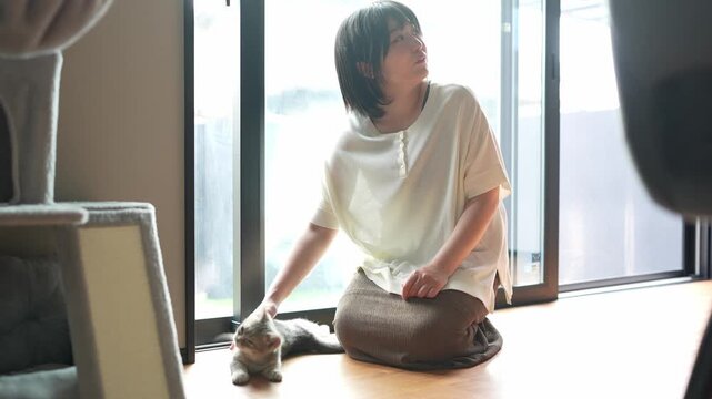 Soft sunlight bathes a quiet room. Woman sits cross-legged near a window. She gently pets a resting cat. Another cat curiously approaches nearby. Peaceful, domestic moment captured indoors