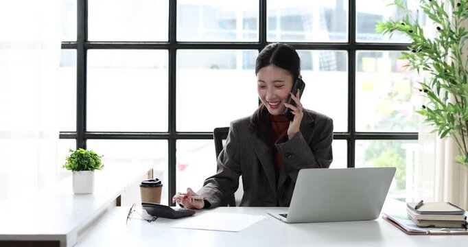Asian businesswoman working in a professional office, multitasking with a laptop and smartphone while reviewing documents, embodying efficiency and modern corporate communication