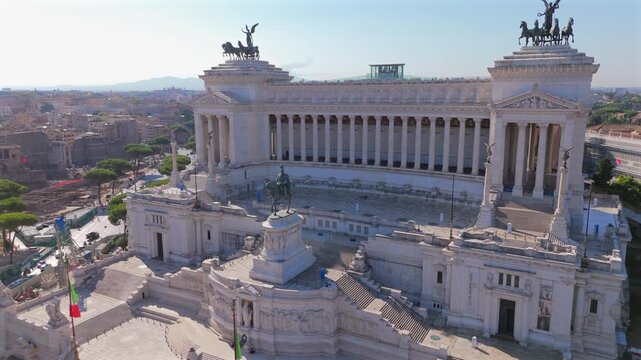 Aerial drone shot in Rome capturing Piazza Venezia and majestic Altare della Patria monument. White marble columns and bronze quadriga statues crown neoclassical architecture against a clear blue sky.