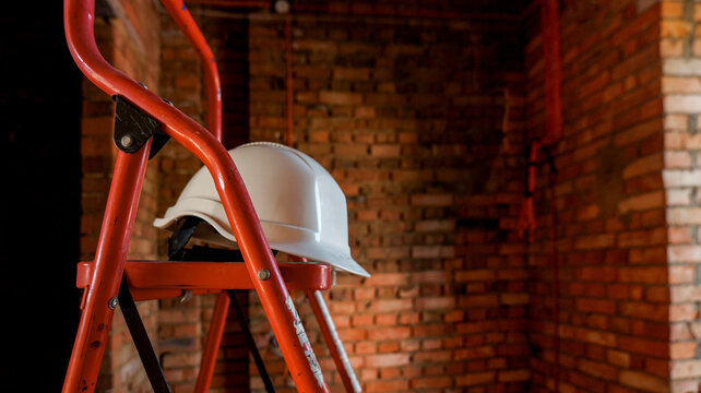 White hard hat resting on a red stepladder against a brick wall background