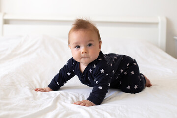 Fototapeta premium A cute little caucasian six-month-old baby boy, dressed in a black jumpsuit with stars, is crawling on all fours on a bed with white linens. Lifestyle close-up portrait in home setting. Real child. 
