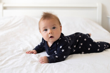 Fototapeta premium A cute little caucasian six-month-old baby boy, dressed in a black jumpsuit with stars, is crawling on all fours on a bed with white linens. Lifestyle close-up portrait in home setting. Real child. 