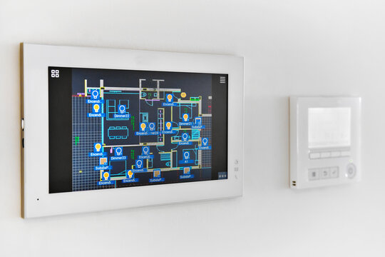 A smart home automation system is mounted on white wall, displaying digital floor plan with various controls, closeup