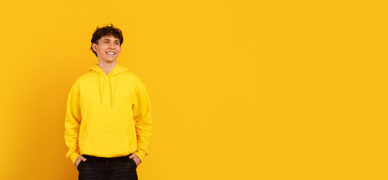 Smiling young man in yellow hoodie looking aside at copy space on yellow studio background, glancing away with relaxed casual confident expression