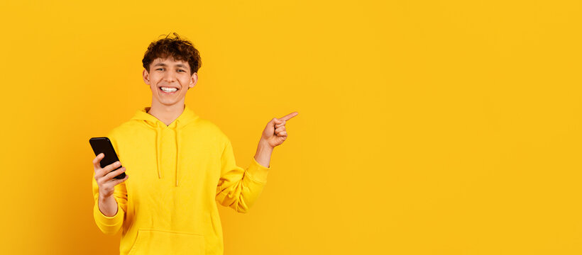 Smiling young guy in yellow hoodie holding smartphone and pointing to empty space on yellow studio background for app, offer or digital promotion, panorama