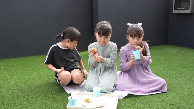 Three children sit on a blanket, eating together. They enjoy simple snacks, sharing moments of joy. The setting is calm and cozy, with green grass and soft lighting. Perfect for family scenes