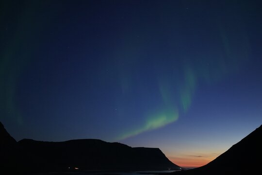 Aerial view of the aurora borealis illuminating the night sky above the dark mountain silhouettes of Flateyri, Isafjardarbaer, Iceland.