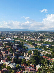 High-angle panoramic daytime cityscape of Tbilisi, Georgia, featuring the Kura River, the Bridge of Peace, and historic old town architecture under a blue sky with clouds © Yafea