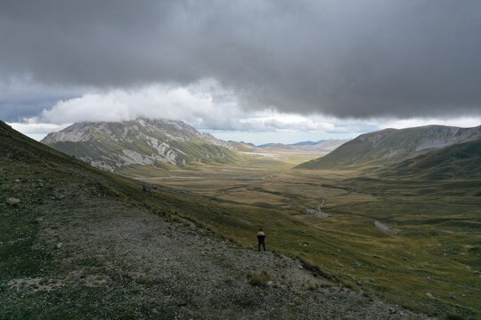 Aerial view of a person standing on a grassy slope overlooking the vast Campo Imperatore plateau with dramatic clouds and rolling mountains in L'Aquila, Abruzzo, Italy.
