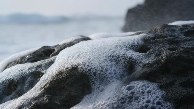 Low angle close view of white sea foam cascading and settling over dark porous lava rock at waterline under soft overcast daylight with textured coastal detail, with foam, bubble, rock, coast detail