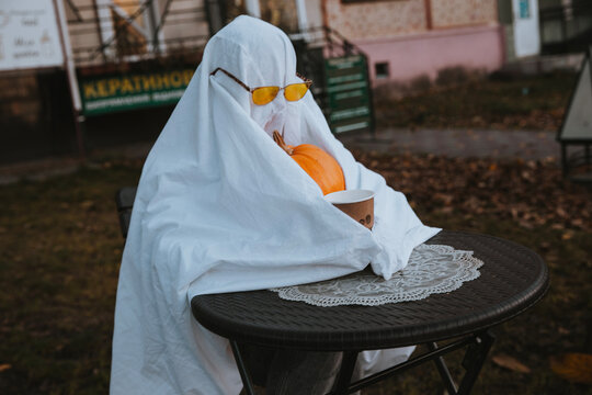 A ghost in sunglasses sitting at a cafe table with a pumpkin and coffee.