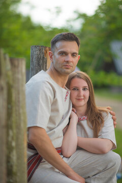 Beautiful young couple in traditional Slavic linen clothing embracing near rustic wooden fence with copy space