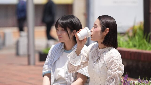Two women sip coffee, smiling gently. Sunlight glows on their casual, friendly expressions. They stroll on a brick-paved sidewalk. Flowers bloom beside a low stone wall