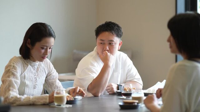 Three people sit at a table in a calm cafe. They sip drinks, chat, and gesture with their hands. The woman on the left smiles gently at her companions. The man in the center rests his chin on his hand