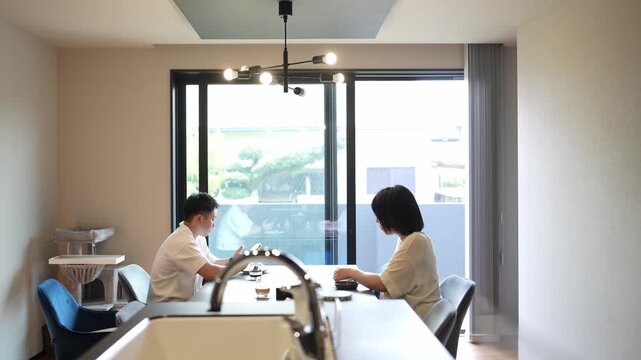 Two people sit at a modern kitchen table. They sip drinks, smiling gently. Natural light fills the minimalist space. The scene evokes calm, domestic comfort. Ideal for lifestyle, interior