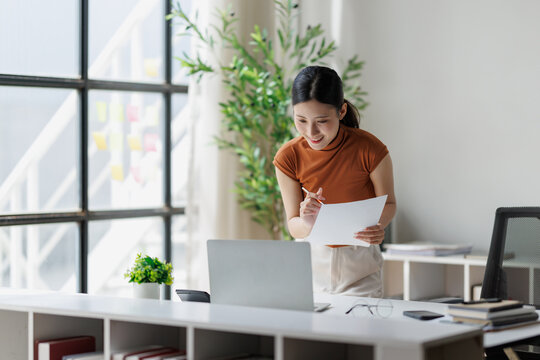 Asian businesswoman analyzing financial data and paperwork, working remotely from a contemporary home office space, staying productive and connected with online tools