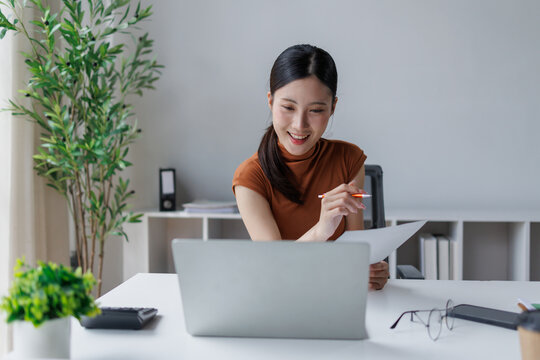 Young asian business professional enjoying remote work, smiling while reading business documents and participating in a virtual online meeting on a laptop