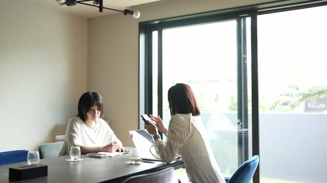 Two women collaborate at a modern conference table. One gestures thoughtfully while the other reviews data. Bright windows frame a calm, professional setting. Ideal for business, education