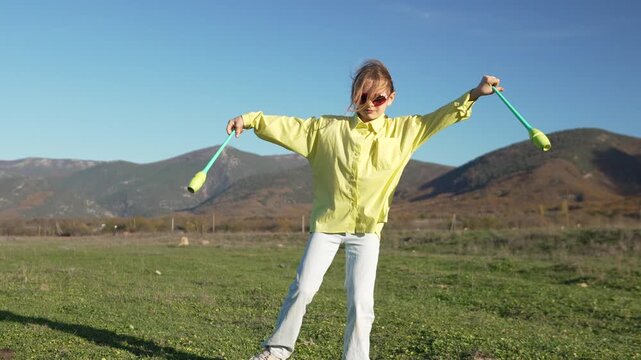 Gymnastics clubs girl practicing exercises outside on grass meadow near scenic mountains in bright sunshine.