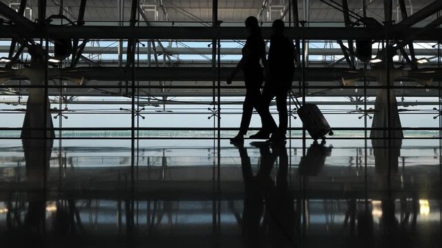 Relaxed passengers couple walks at terminal, stop to look to airport apron. Silhouetted shot at dim concourse, two people spend time before boarding starts, stroll around modern terminal