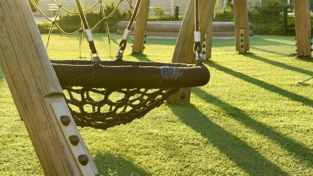 A large rope nest swing sits peacefully in the golden evening light on a lush green lawn. This calm scene brings a feeling of quiet rest and nostalgia to the empty neighborhood park.
